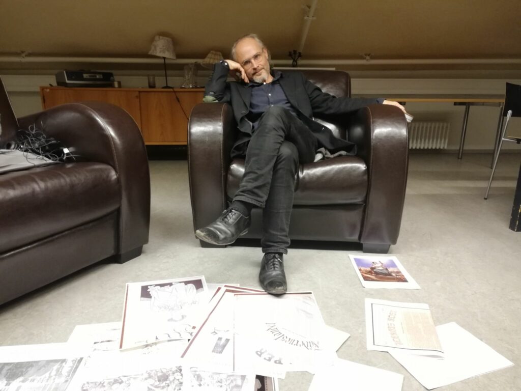 Fred Andersson sitter i en fåtölj med teckningar utbredda på golvet framför sig.