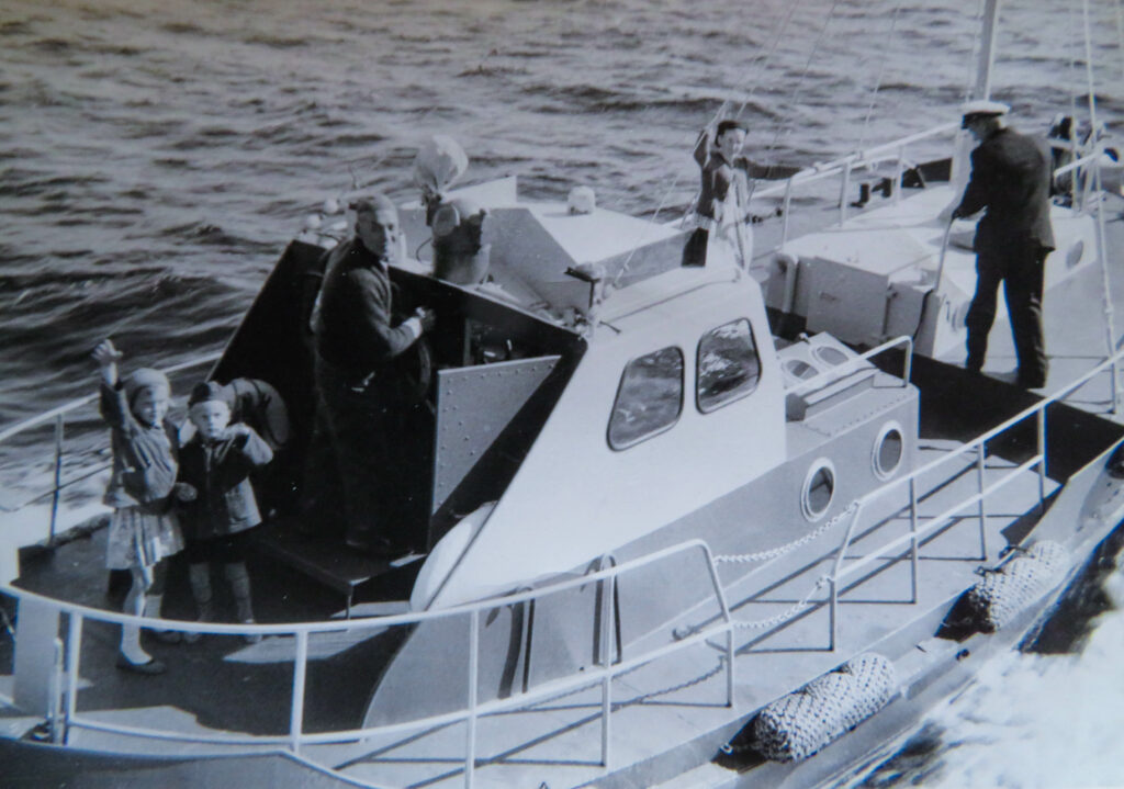 Gammal svartvit bild på någon som styr en mindre båt på havet med några passagerare.