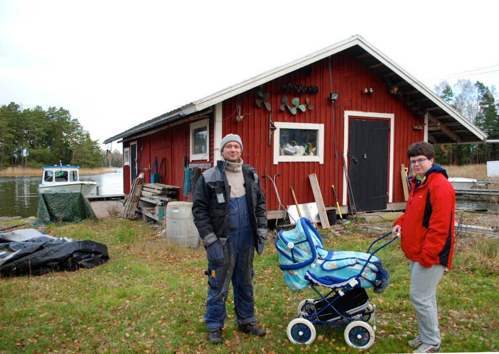 Ett par med barnvagn framför ett rött hus och en liten brygga.